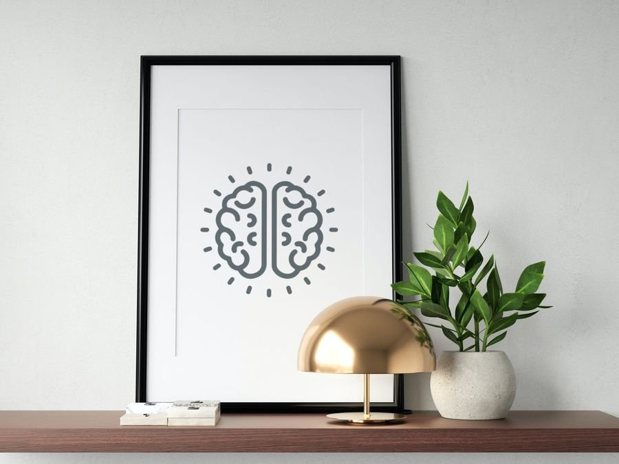 The brain mark in a black frame on a wooden shelf beside a brass lamp and a plant